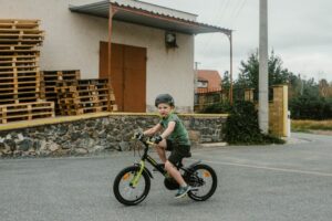 A young boy in a helmet rides his bicycle near a warehouse with stacked wooden pallets.