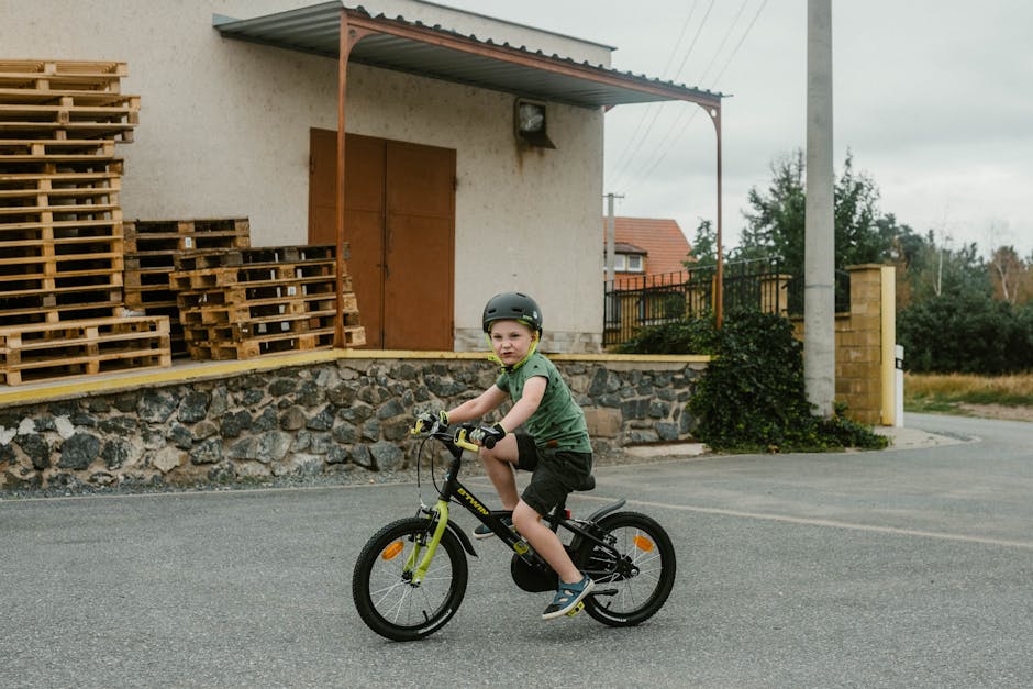 A young boy in a helmet rides his bicycle near a warehouse with stacked wooden pallets.
