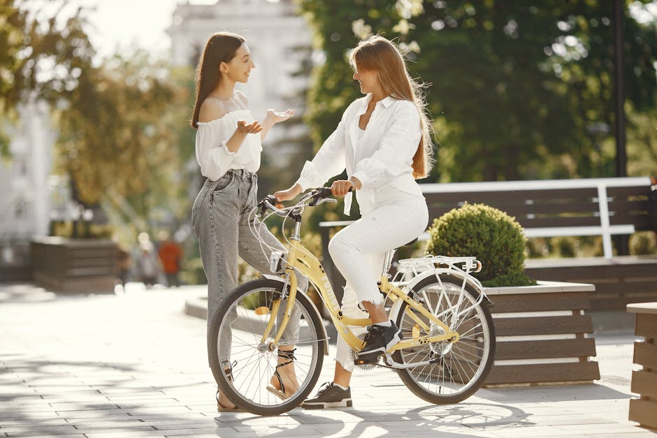 Two women having a conversation, one standing and the other seated on a yellow bicycle in an urban park setting.