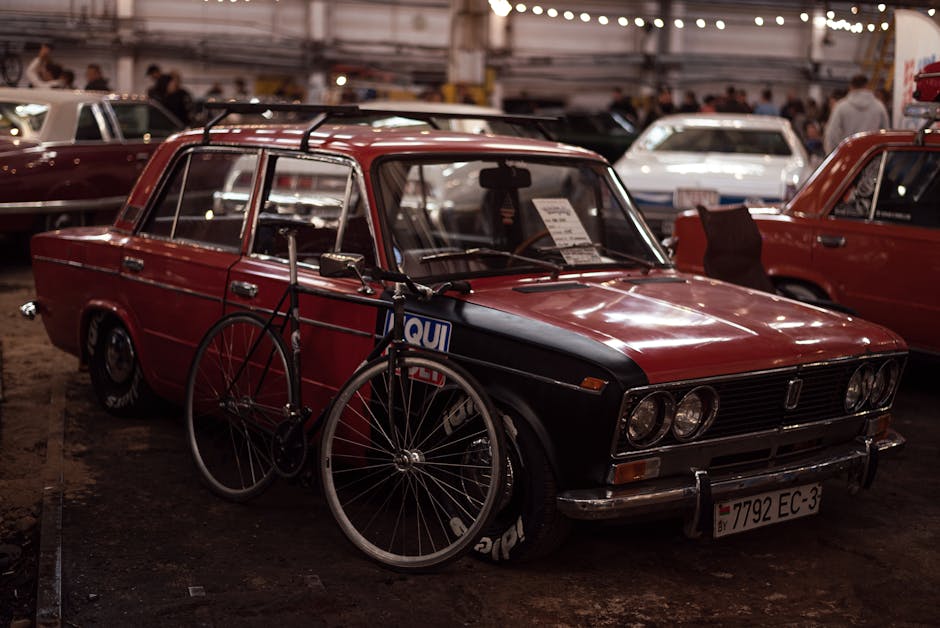Vintage red car with a bicycle leaning against it at an indoor car exhibition event.