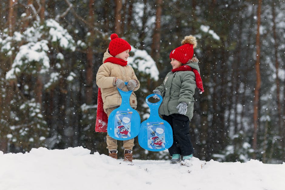 Two kids in winter attire holding sleds, enjoying a snowy day outdoors.