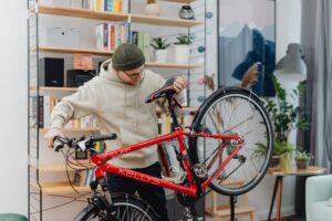 A man in a hoodie repairs a red bicycle in a cozy indoor setting with shelves and plants.
